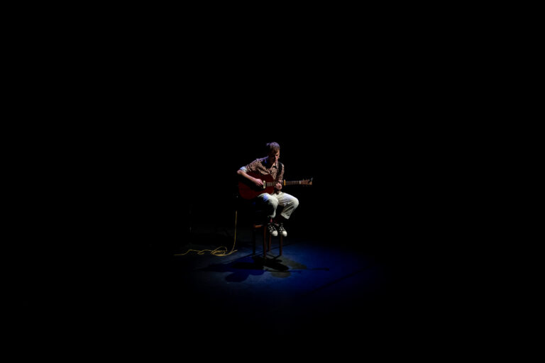 Een muzikant zit op een stoel in een bijna volledig donkere setting en speelt gitaar onder een blauwe lichtstraal.