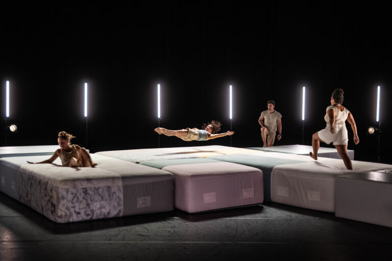 Een groep dansers in beweging op grote matrassen onder fel licht in een donkere ruimte.