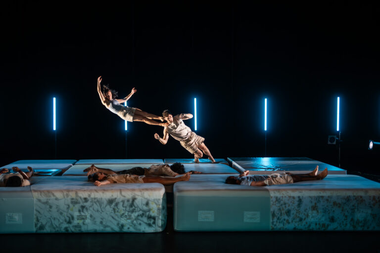 Twee acrobaten springen en zweven boven een reeks matrassen, terwijl andere performers op de matrassen liggen in een donkere ruimte met blauwe verlichting.