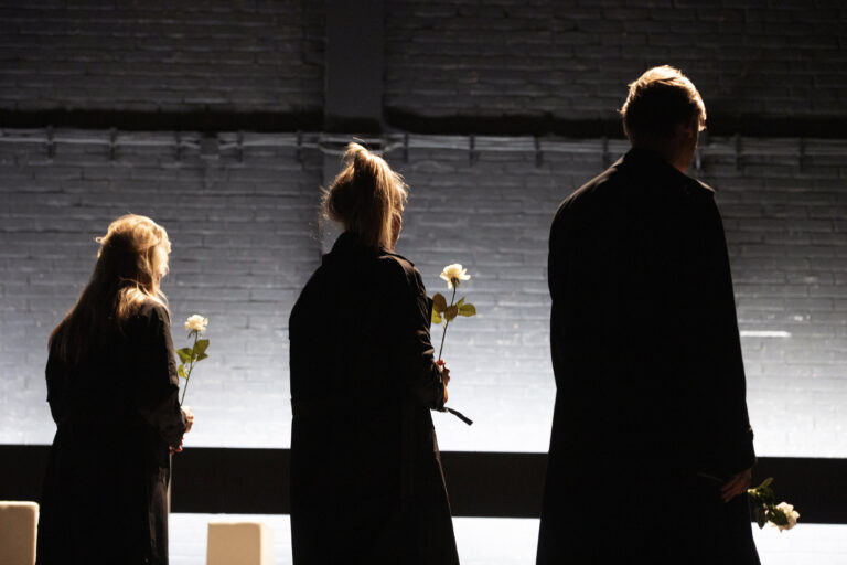 Drie personen als donkere silhouetten staan met hun ruggen naar de camera tegen een fel verlichte achtergrond.