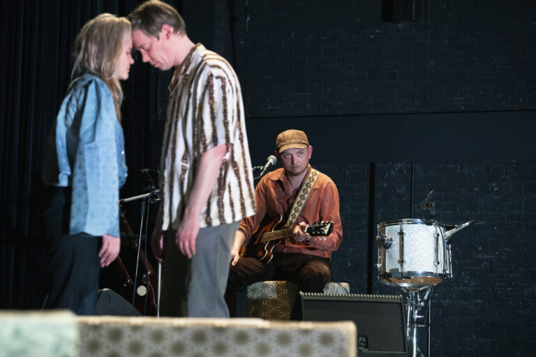 Een gitarist zit op een stoel en speelt gitaar op een podium, met een drummer en drumstel op de achtergrond.