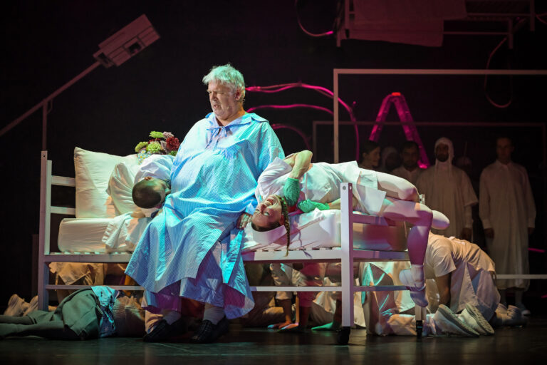 Een acteur in een helder blauw kostuum zit op een bank op een theatertoneel, met andere acteurs op de achtergrond.