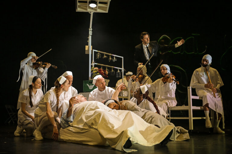 Een groep mannen in lange, lichte gewaden zit op het podium tijdens een toneelvoorstelling.