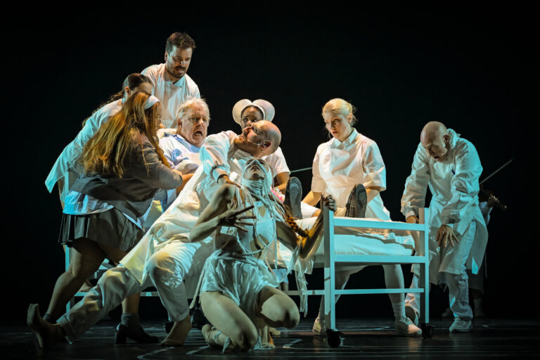 Meerdere acteurs in witte kleren staan en zitten op een donker podium rondom een persoon die op de grond ligt naast een witte stoel.