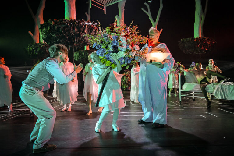 Een jonge danseres in een witte jurk danst op een donker podium, met een kleurrijk bloemenarrangement op de achtergrond.