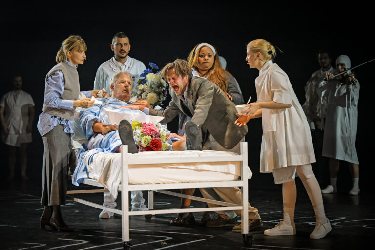 Theater-scène met een patiënt liggend in een ziekenhuisbed, omringd door meerdere acteurs.