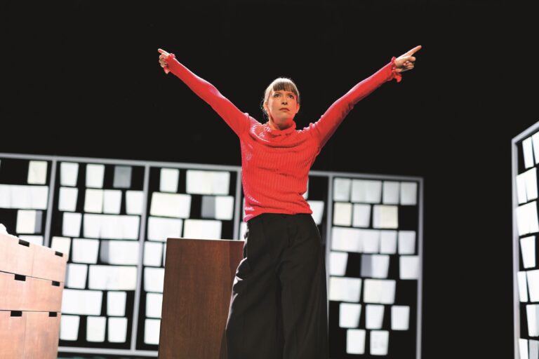 Actrice Maria Kraakman staat met een rode trui en zwarte rok met haar armen wijd in een kamer met houten kasten.