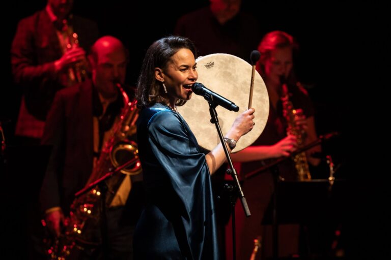 Charly Lowry, een native American zangeres, zingt en bespelt een grote handtrommel. Op de achtergrond enkele blazers