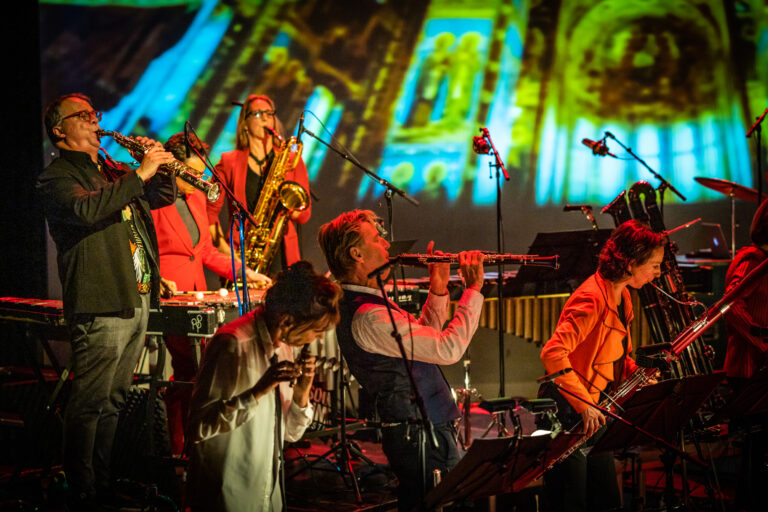 Zes musici van het Nederlands Blazers Ensemble in actie, tegen het decor van een sfeervol verlichte kerk.