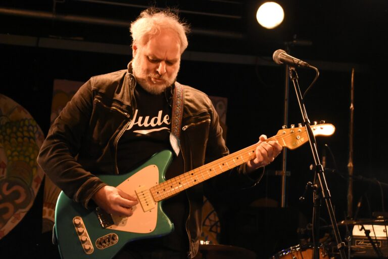 Foto van een oudere man met wit haar die een unieke gitaar bespeelt op een podium, met een spotlight achter hem.