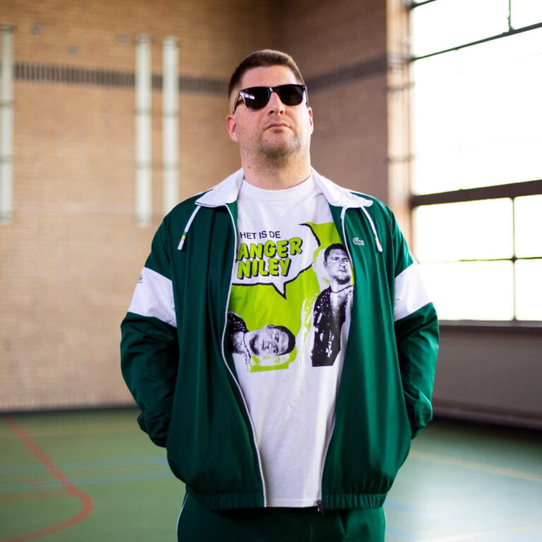 Een man in een groen sportjack en zonnebril poseert in een gymzaal.
