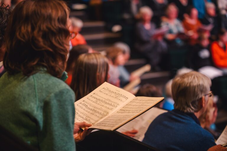 Een persoon met bruin haar houdt een muziekpartituur vast, terwijl een publiek in de achtergrond aan het luisteren is.