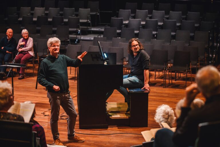 Twee mannen staan naast een grote vleugel op een leeg podium in een concertzaal.
