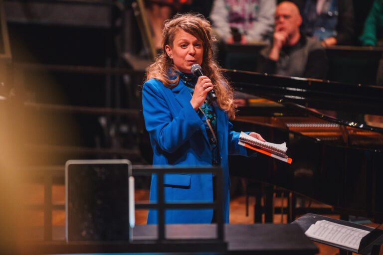 Een vrouw in een blauwe blazer zingt in een microfoon op een podium, met een piano en muzikanten op de achtergrond.