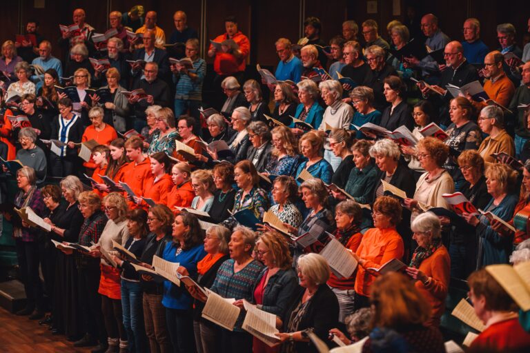 Een groot koor van mensen in verschillende kleding, zingend en bladmuziek houdend in een zaal.