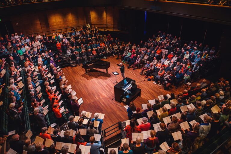 Een groot gezelschap van zangers en een begeleidende piano in een theateromgeving.