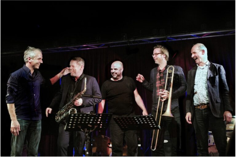 Een groep muzikanten staat lachend op het podium na een optreden, met onder andere een saxofoon en een trombone zichtbaar.