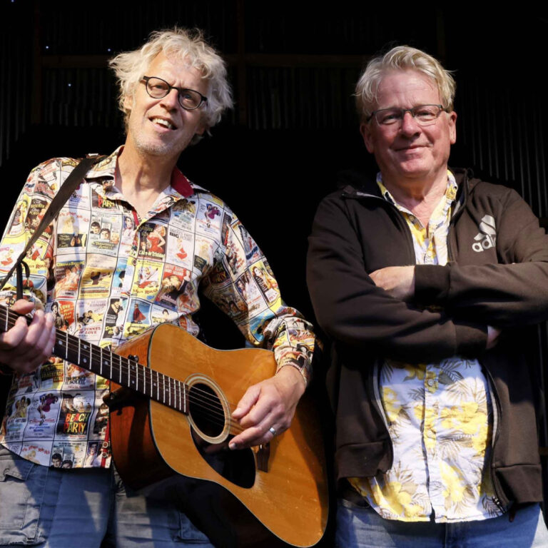 Twee mannen, Pe en Rinus, staan naast elkaar, waarvan één een gitaar vasthoudt en beiden kleurrijke overhemden dragen