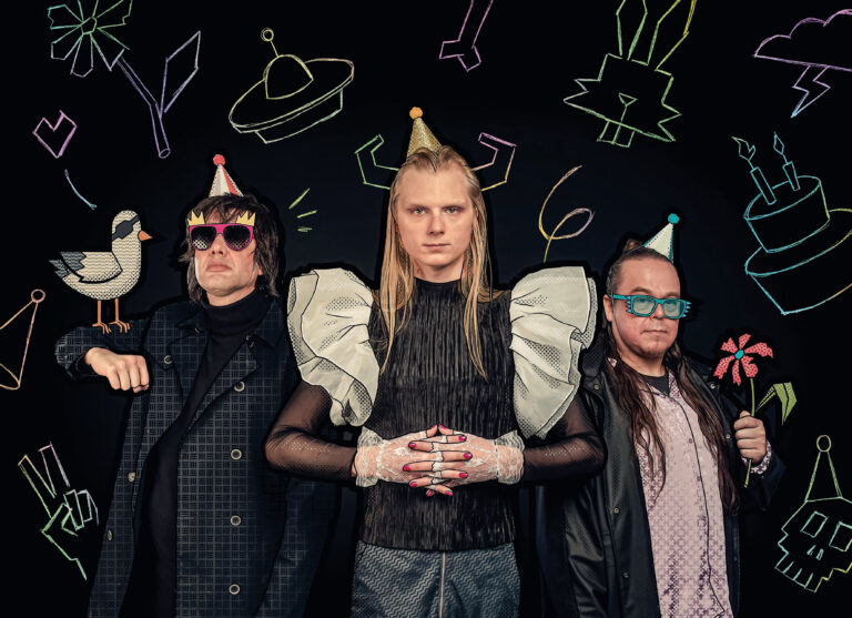 Een groepsfoto van drie mensen tegen een zwarte achtergrond met kleurrijke neon krijtachtige tekeningen van feestartikelen, zoals een hoed, een eend, en bliksemschichten