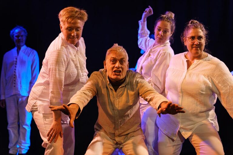 Centraal staande danser die naar voren reikt, omringd door andere dansers op een fel verlicht podium.