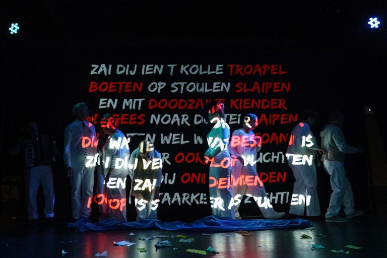 Een scène met acteurs in witte kleding, waarop krachtige teksten in rode en witte letters worden geprojecteerd, wat een emotioneel en dramatisch effect creëert tegen een donkere achtergrond, terwijl de acteurs stilstaan in contemplatieve houdingen.