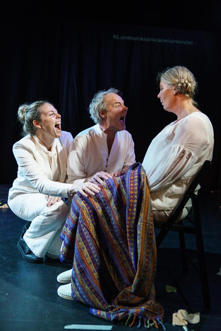 Een intense scène waarin twee acteurs in witte kleding expressief schreeuwen richting een derde acteur die stil en met een serieuze blik op een stoel zit, met een kleurrijke deken over haar schoot, tegen een donkere achtergrond die de dramatische spanning benadrukt.