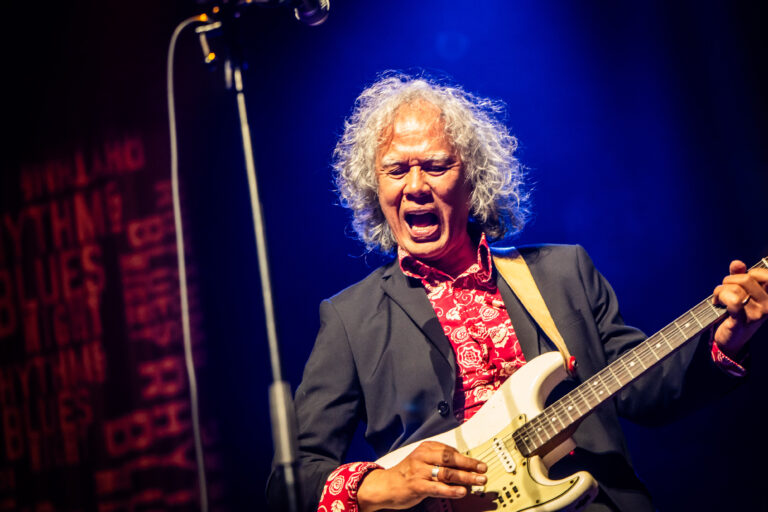 Een performer met krullend wit haar speelt gitaar en zingt op een podium met blauwe verlichting.