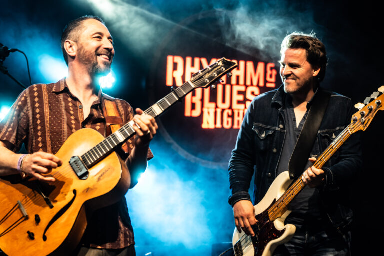 Twee muzikanten voeren een live optreden uit op een podium, met een achtergrond van rook en een verlichte tekst