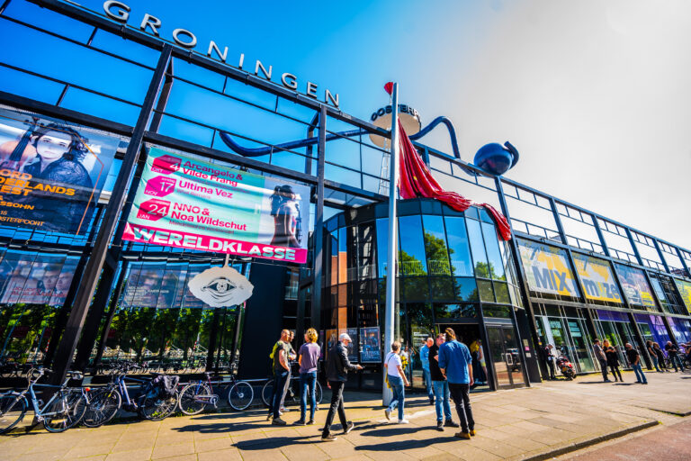 Een groep mensen staat voor een modern gebouw met grote ramen en een vlag, onder een heldere blauwe lucht.
