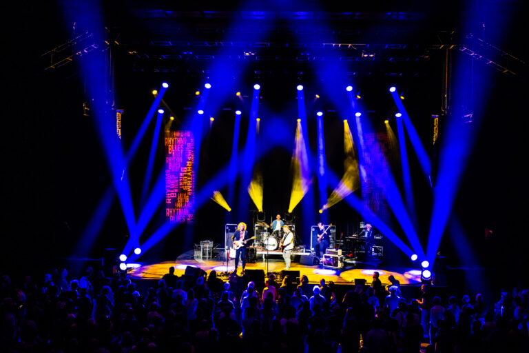 Een live band optredend op een verlichte stage met kleurrijke lichtstralen en een enthousiast publiek.
