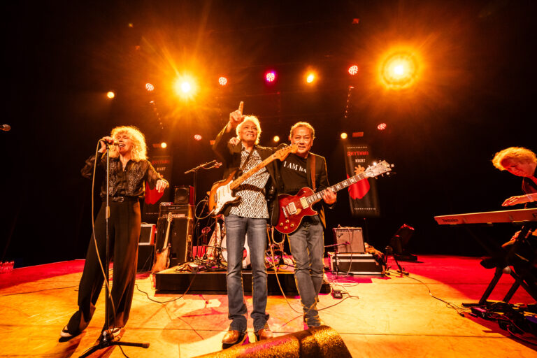 Een band staat op het podium en speelt muziek onder felgekleurde lichten.