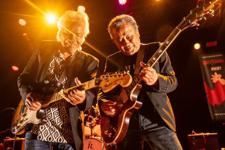 Twee muzikanten spelen samen op het podium met elektrische gitaren, omringd door fel licht.