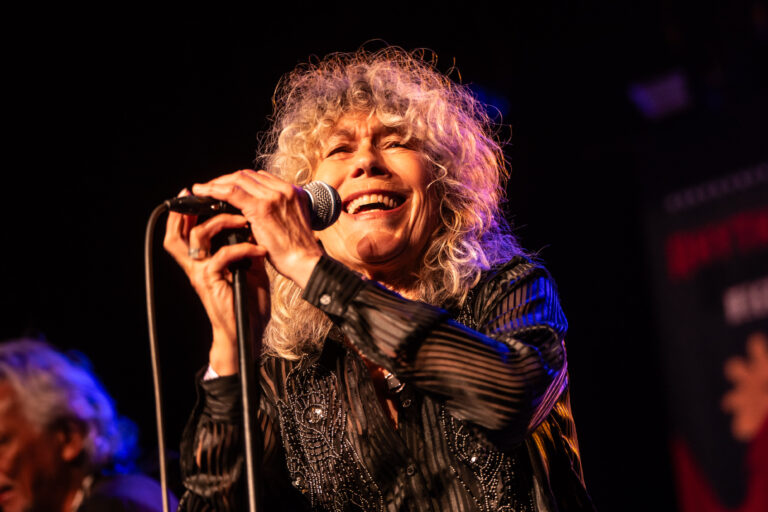 Een zangeres met een krullende, blonde haardos houdt een microfoon vast en lacht terwijl ze optreedt op het podium.