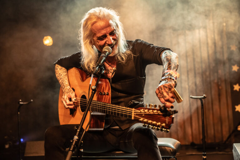 Een muzikant met lange, grijze haren speelt een akoestische gitaar terwijl hij zingt op een podium met rokerige verlichting.