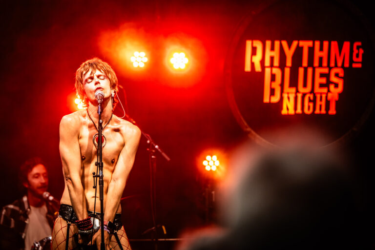 Een performer zingt op een podium, verlicht door oranje lichten, met een spandoek waarop 