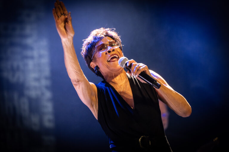 Een zangeres staat op het podium en zingt met een microfoon, terwijl ze haar hand omhoog steekt.