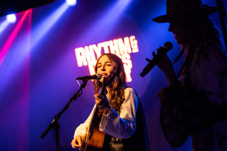 Een vrouwelijke muzikant speelt gitaar en zingt op een podium met een fel verlichte achtergrond.