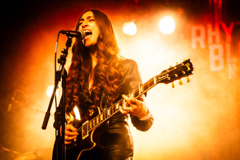 Een muzikante met lange haren speelt gitaar en zingt op een podium met warme verlichting.