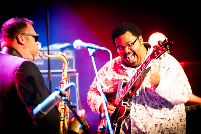 Twee muzikanten geven een energiek optreden, waarbij een man op een gitaar speelt en een andere man met een saxofoon naast hem staat.