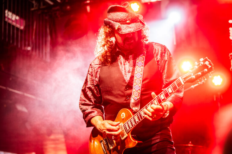 Een muzikant speelt gitaar op een podium met felrode verlichting en rookeffecten.