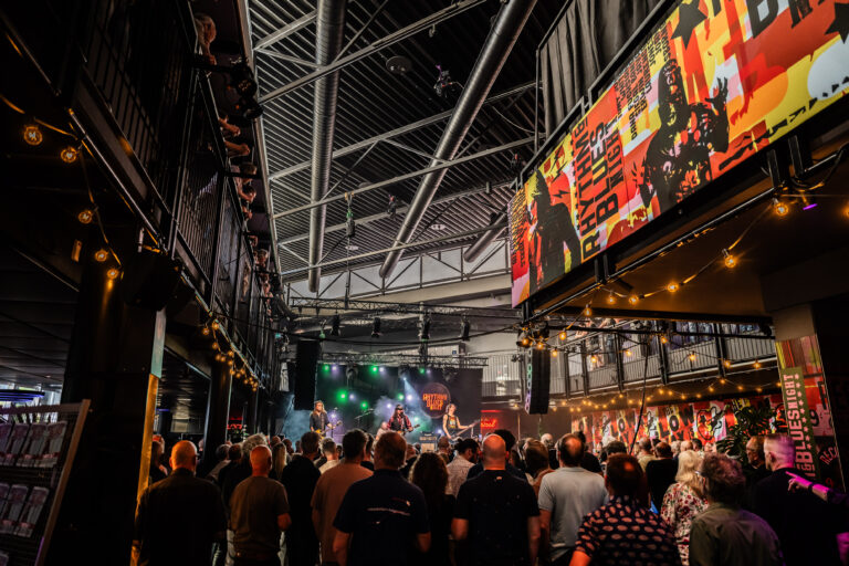 Een drukbezocht concert met een levendige sfeer en een groot scherm dat beelden toont.