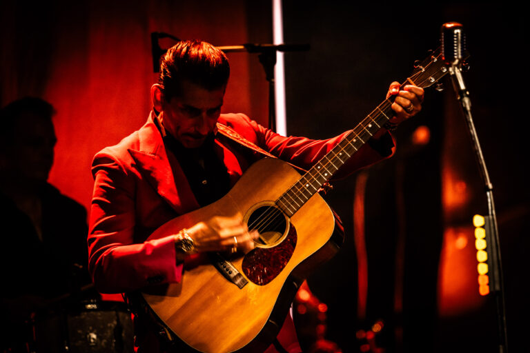 Een muzikant in een felrood pak staat op het podium en speelt op een akoestische gitaar.