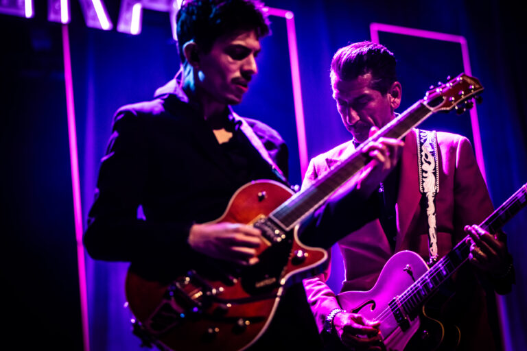 Twee muzikanten spelen gitaar op een podium met kleurrijke verlichting.