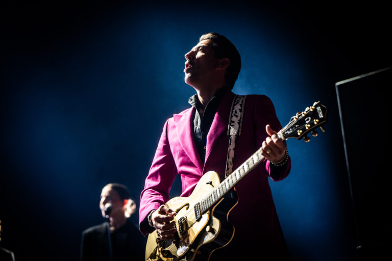 Een gitarist in een opvallende roze jas speelt een elektrische gitaar op een podium, terwijl een andere muzikant op de achtergrond zichtbaar is.