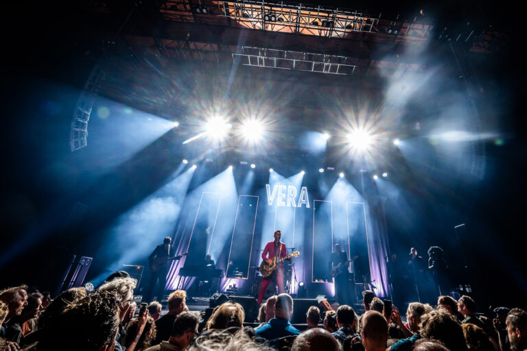 Een muzikant staat op het podium met krachtige verlichting en een enthousiaste menigte voor zich.