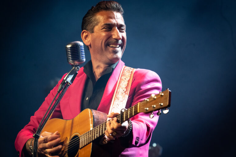 Een muzikant in een felroze kostuum speelt gitaar en zingt op een podium.