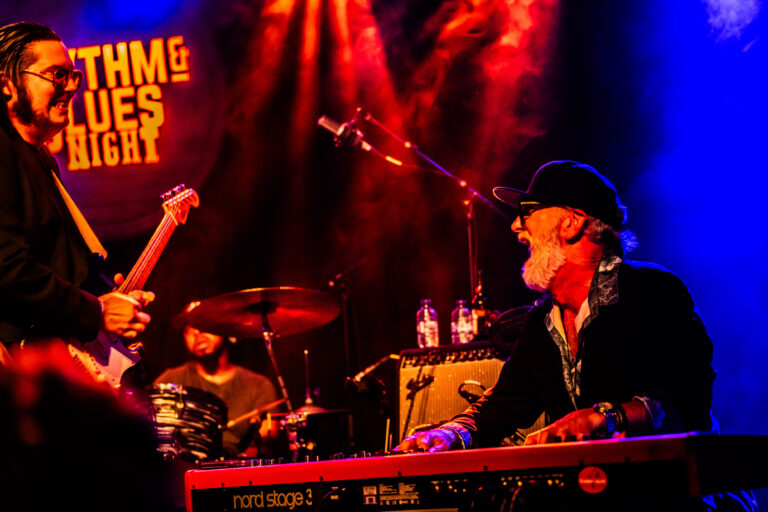 Een muzikant speelt op een keyboard terwijl een andere muzikant op drums speelt, met kleurrijke verlichting op de achtergrond.