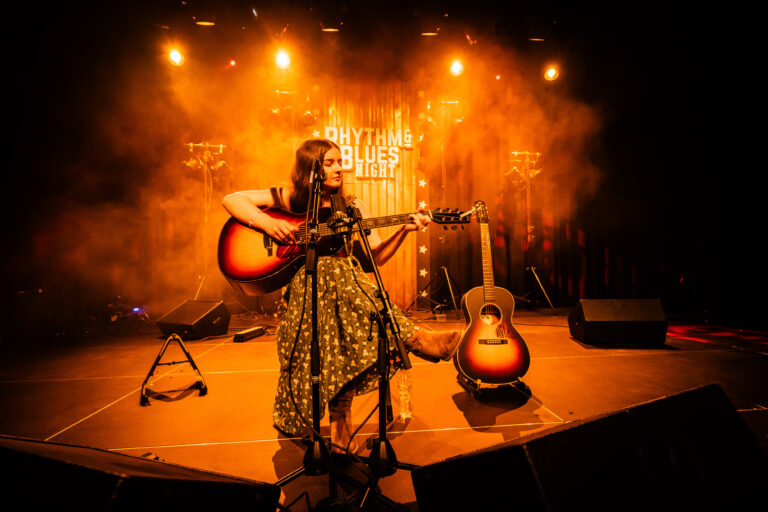 Een muzikante in een groene jurk speelt gitaar op een sfeervol verlichte podium met rook.