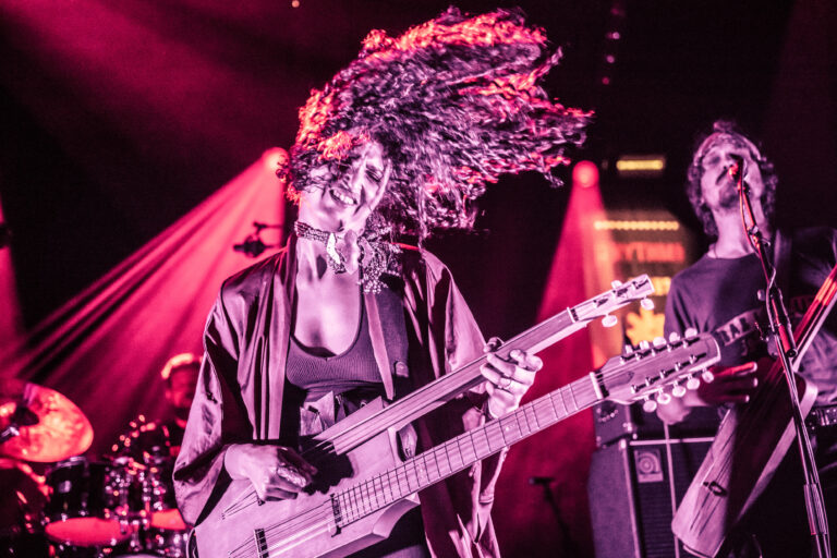 Een muzikante speelt een basgitaar terwijl ze energiek beweegt op het podium, omringd door kleurrijke lichten.