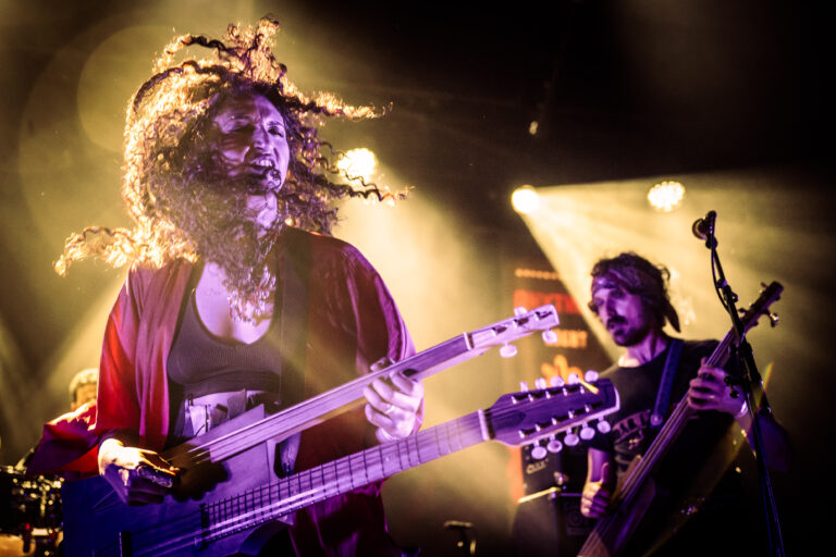 Een muzikant speelt een dubbele gitaar op een podium met verlichting, terwijl een andere muzikant op de achtergrond kijkt.